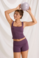 Model holding a Pilates ball overhead in a plum lace-trim tank and matching workout shorts